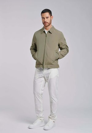 Uomo con capelli corti e barba che indossa una giacca verde oliva con cerniera, pantaloni bianchi e scarpe da ginnastica bianche, in piedi con le mani nelle tasche.