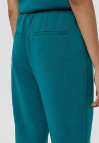 Teal loungebroeken met een gladde textuur, elastische tailleband en twee achterzakken. Het ontwerp is eenvoudig zonder zichtbare patronen of hardware.