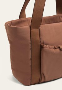 Bruine gestikte tote bag met brede stoffen handvatten, een zijvak en een gladde textuur, met een gestructureerde vorm en een minimalistisch ontwerp.