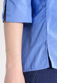 Camicia di cotone azzurro chiaro con maniche arrotolate, spacchi laterali e una texture liscia, parzialmente visibile su un tessuto di jeans scuro.