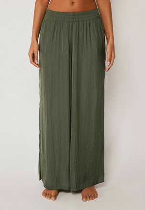 Barfodet person iført løstsiddende, højtaljede olivengrønne bukser med vide ben og elastisk talje, stående mod en ensfarvet baggrund.