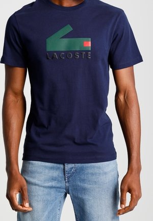 Homme portant un t-shirt Lacoste bleu marine avec un logo vert et rouge et un jean bleu clair, debout les bras détendus le long du corps.