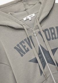 Harmaa vetoketjullinen huppari, joka on valmistettu pehmeästä materiaalista. Edessä on suuri "NEW YORK" -painatus sekä tähtikuvio, ja siinä on säädettävät nyörit.