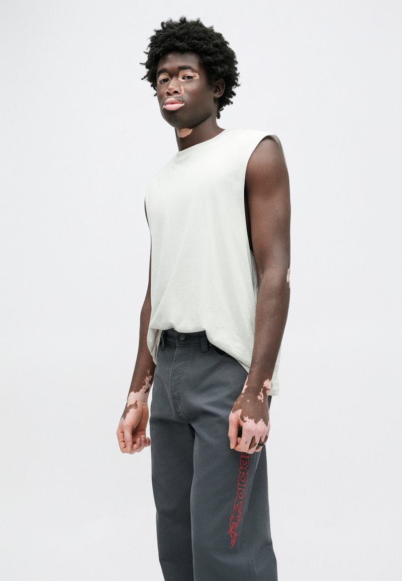 Sleeveless light gray cotton top paired with dark gray jeans featuring a red graphic print on the left leg. Casual fit, smooth texture.