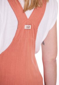 Rostfarbene Leinen-Overalls mit einem Etikett auf der Rückseite, breiten Trägern und einem lässigen Schnitt, kombiniert mit einem weißen T-Shirt.