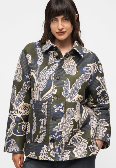 Γυναίκα φοράει ένα κουμπωτό μπουφάν με πράσινο, μπλε και μπεζ λουλουδένιο μοτίβο με μεγάλες τσέπες και μακριά μανίκια, ποζάροντας με το ένα χέρι στην τσέπη.