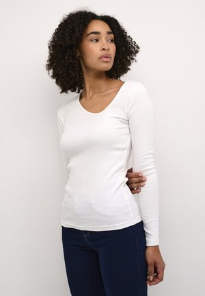 Top bianco a maniche lunghe con texture a costine e scollatura a barchetta, abbinato a jeans blu scuro. Design semplice, silhouette aderente.