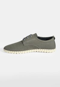 Chaussures perforées vert foncé avec des lacets gris, au design épuré et semelle tissée de couleur claire. Texture lisse et forme basse.
