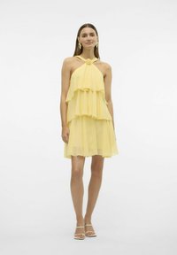 Gul chiffon-kjole med lagdelte volanger, halterneck-design og knyttet frontdetalje. Knælængde med en svævende, let struktur.