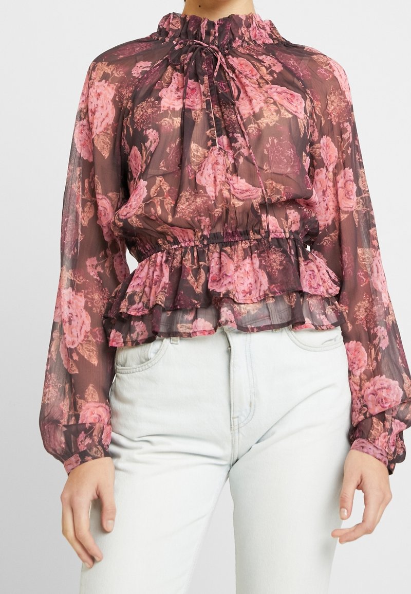 Blouse transparente à manches longues avec motif floral rose, col haut et taille froncés, portée avec un jean bleu clair.