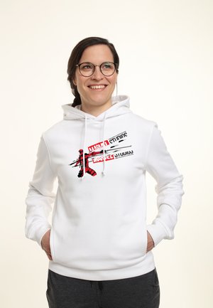 Smilende kvinde iført briller og en hvid hættetrøje med et rødt og sort Deadpool-grafik og Marvel Studios-tekst, hænderne i lommerne.
