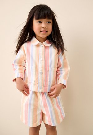 Junges Mädchen mit glatten dunklen Haaren, das eine pastellfarbene gestreifte Jacke und passende Shorts vor einem einfarbigen hellen Hintergrund trägt.