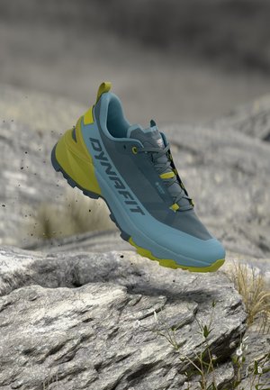 Blauw-gele Dynafit trailrunningschoen die boven rotsachtig terrein met kleine planten en een vage achtergrond in de lucht zweeft.