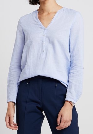 Femme portant un chemisier bleu clair à manches longues et col en V, ainsi qu'un pantalon bleu marine, avec une montre marron au poignet gauche, debout devant un fond uni.