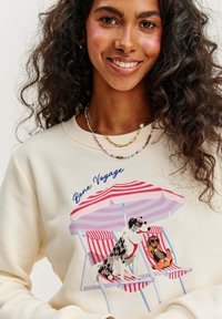 Krémová mikina s ilustrací dvou psů pod červenobílým pruhovaným deštníkem, s nápisem "Bone Voyage."