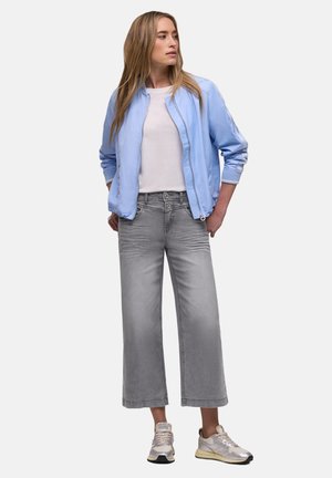 Donna in piedi con le mani nelle tasche, indossa una giacca bomber azzurro chiaro, una t-shirt bianca, jeans grigi a gamba larga e sneaker bianche.