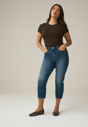 Woman standing with hands in pockets wearing a fitted dark brown short-sleeve shirt, cropped blue jeans, and snake-print flats.