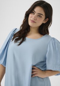 Blouse bleu clair avec des manches bouffantes, encolure ronde et texture lisse. Tombe avec souplesse pour un ajustement décontracté. Pas de motifs ni d'accents visibles.
