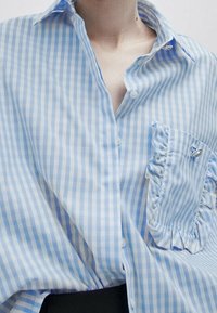 Camicia a righe blu e bianche con taschino arricciato, design con bottoni e un piccolo emblema decorativo sul colletto. Tessuto di cotone.