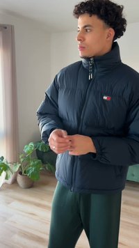 Μαύρο μπουφάν τύπου puffer με ψηλό γιακά, κλείσιμο με φερμουάρ και λεπτομέρειες λογότυπου. Μαλακή, επενδυμένη υφή, φορεμένο πάνω από σκούρο πράσινο παντελόνι. Εσωτερικός χώρος.