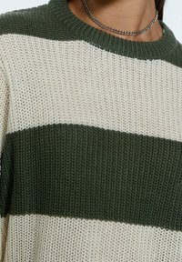 Knitted sweater with green and cream horizontal stripes. Features a ribbed neckline and loose weave texture. Silver chain necklace visible.