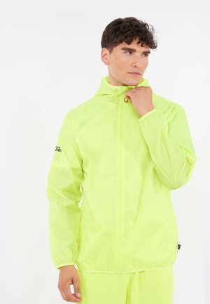 Jonge man die de kraag van een neongele lichtgewicht jas met een voorrits aanpast, staande tegen een effen witte achtergrond.