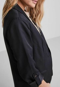 Námořnický modrý blazer s upraveným střihem, dlouhými rukávy, klopovým límcem a knoflíkovými detaily na manžetách. Hladká textura látky.