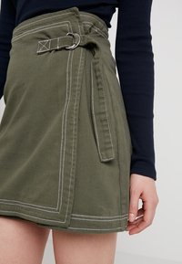 Falda envolvente verde oliva hecha de un tejido resistente, con costuras blancas, un lazo para el cinturón con un anillo plateado y un detalle de lazo lateral.
