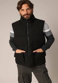 Zwarte gewatteerde gilet met opstaande kraag, blinde rits aan de voorkant en twee zijzakken. Gedragen over een grijs shirt met lange mouwen en gestreepte accenten.