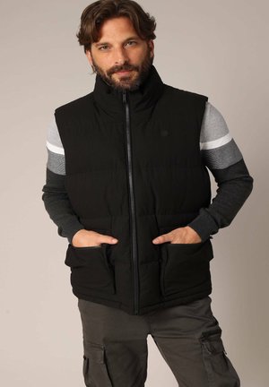 Gilet rembourré noir avec col montant, fermeture éclair sur le devant et deux poches latérales. Porté par-dessus un t-shirt à manches longues gris avec des accents rayés.