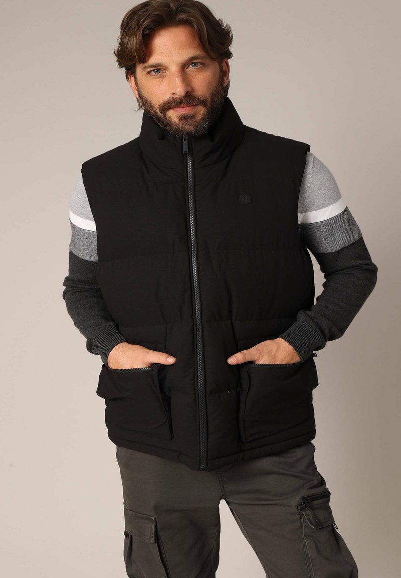 Gilet rembourré noir avec col montant, fermeture éclair sur le devant et deux poches latérales. Porté par-dessus un t-shirt à manches longues gris avec des accents rayés.
