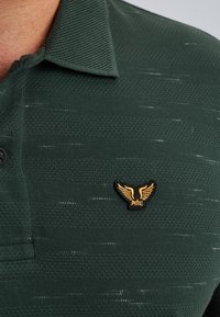 Camisa polo de color verde oscuro con un tejido texturizado y un patrón sutil, un solo botón negro y un logo dorado con alas en el pecho.