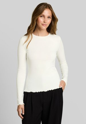 Femme aux longs cheveux châtain clair portant un haut blanc à manches longues et un pantalon noir, debout avec une main dans la poche sur un fond gris.