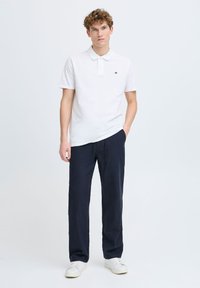 White polo shirt with a collar and logo, paired with navy drawstring trousers and white sneakers, against a plain background.