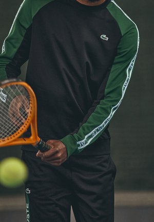 Osoba ubrana w czarne i zielone sportowe ubrania Lacoste, trzymająca pomarańczową rakietę tenisową, przygotowująca się do uderzenia żółtej piłki tenisowej.