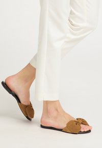 Infradito in suede marrone con dettaglio a fiocco, con una suola nera piatta. Pantaloni bianchi parzialmente visibili, che evidenziano le calzature.
