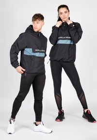 Sort hættetrøjer med lyseblå horisontale striber, kombineret med sorte leggings og kontrasterende sportssko. Glat stof, funktionelt design.
