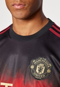 Jersey de Manchester United con degradado negro y rojo, que presenta un collar redondo, tres rayas rojas en los hombros y un logo bordado en dorado.
