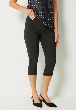 Femme portant des leggings courts noirs, des talons slingback noirs et un haut sans manches bleu marine à pois avec bordure en dentelle.