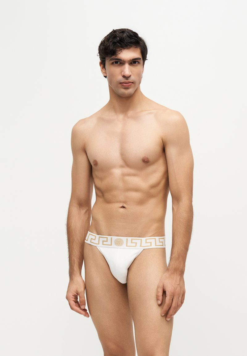 Jeune homme aux cheveux foncés portant un slip blanc avec une ceinture décorative dorée, debout devant un fond clair uni.