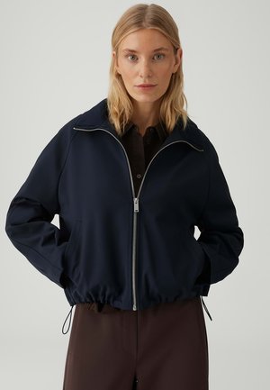 Donna bionda che indossa una giacca blu navy con cerniera e orlo elastico, con le mani nelle tasche, abbinata a pantaloni marrone scuro, sfondo neutro.