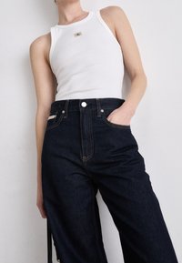 Weißes, geripptes Tanktop mit einem Logopatch; dunkelblaue, hochtaillierte Jeans mit geradem Bein und sichtbaren Steppnähten.