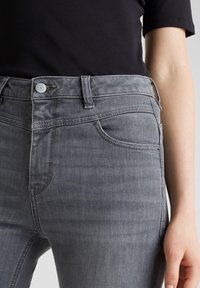 Grå denimjeans med en figurnära design, femfickorsstil och en synlig knappstängning vid midjan. Slät textur och lätt stretch.