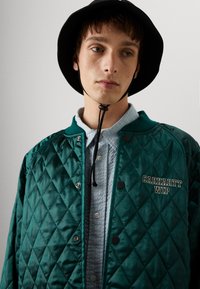 Chaqueta bomber acolchada verde con botones negros y logo "Carhartt WIP". Superpuesta sobre una camisa texturizada azul claro. Sombrero bucket negro.