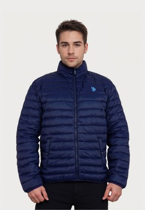 Mladý muž v tmavě modré prošívané bundě na zip s malým světle modrým logem na hrudi, stojící před jednoduchým pozadím.