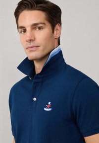 Polo shirt en coton navy avec un col structuré, présentant une doublure intérieure rayée bleue et un logo brodé sur la poitrine.
