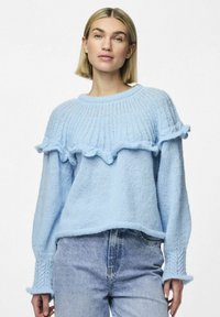  svetlo modra pletena pulover s nabrano obrobo, širokimi manšeti in teksturiranim vzorcem. V kompletu z svetlo modrimi kavbojkami.