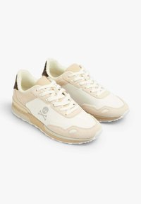 Baskets beiges avec une semelle intermédiaire texturée, une tige en cuir lisse, un logo de crâne, et des accents scintillants sur le talon. Elles disposent de lacets et d'une doublure blanche.