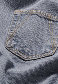 Denim stof met een grote, schuine patch zak met contrasterend gele stitching. Gladde textuur met een vervaagde blauwe kleur.