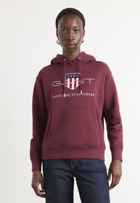 Felpa bordeaux realizzata in tessuto morbido, dotata di tasca frontale, cappuccio con cordino e logo a scudo stampato con dettagli "GANT" e "1949".